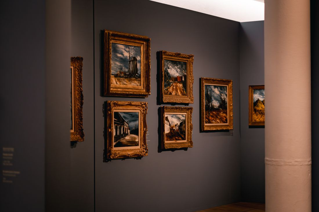 A dimly lit gallery room with several framed paintings hung along a dark wall