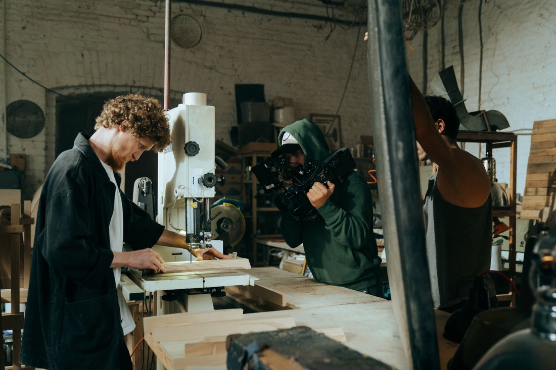 A camera operator films a woodworker guiding a plank through a bandsaw in a workshop, the operator's face turned toward the camera viewfinder rather than the lens