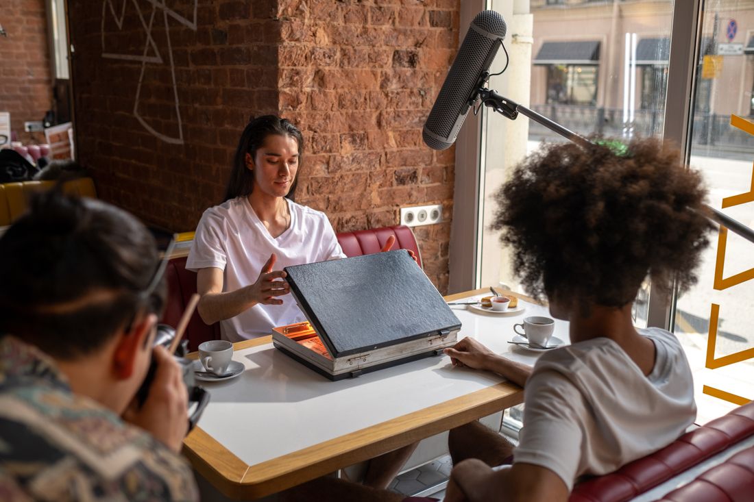 A boom microphone extends over a cafe table where a woman gestures while talking to two people whose backs face the viewer, a window with a street view behind them