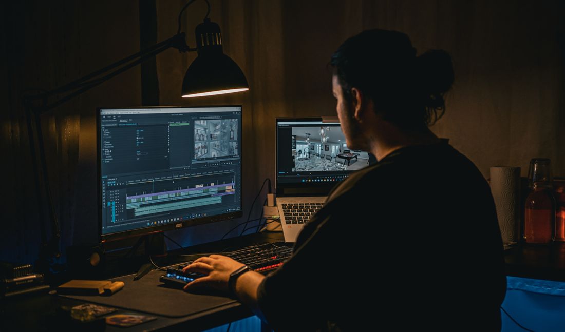 A person seen from behind sits at a desk editing video, one hand on the keyboard, a monitor showing an editing timeline with an interior scene and no faces visible, the room lit mostly by screen glow