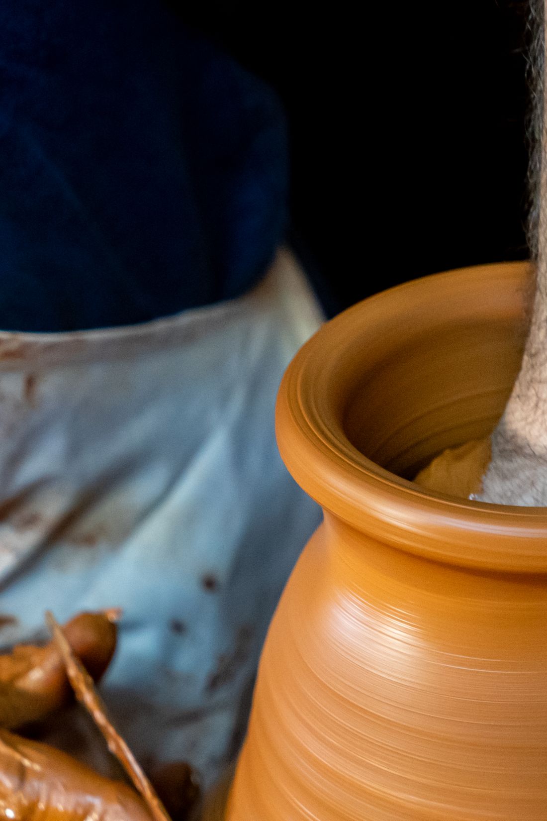 Close crop of hands shaping wet clay on a pottery wheel, textured orange clay against a dark apron