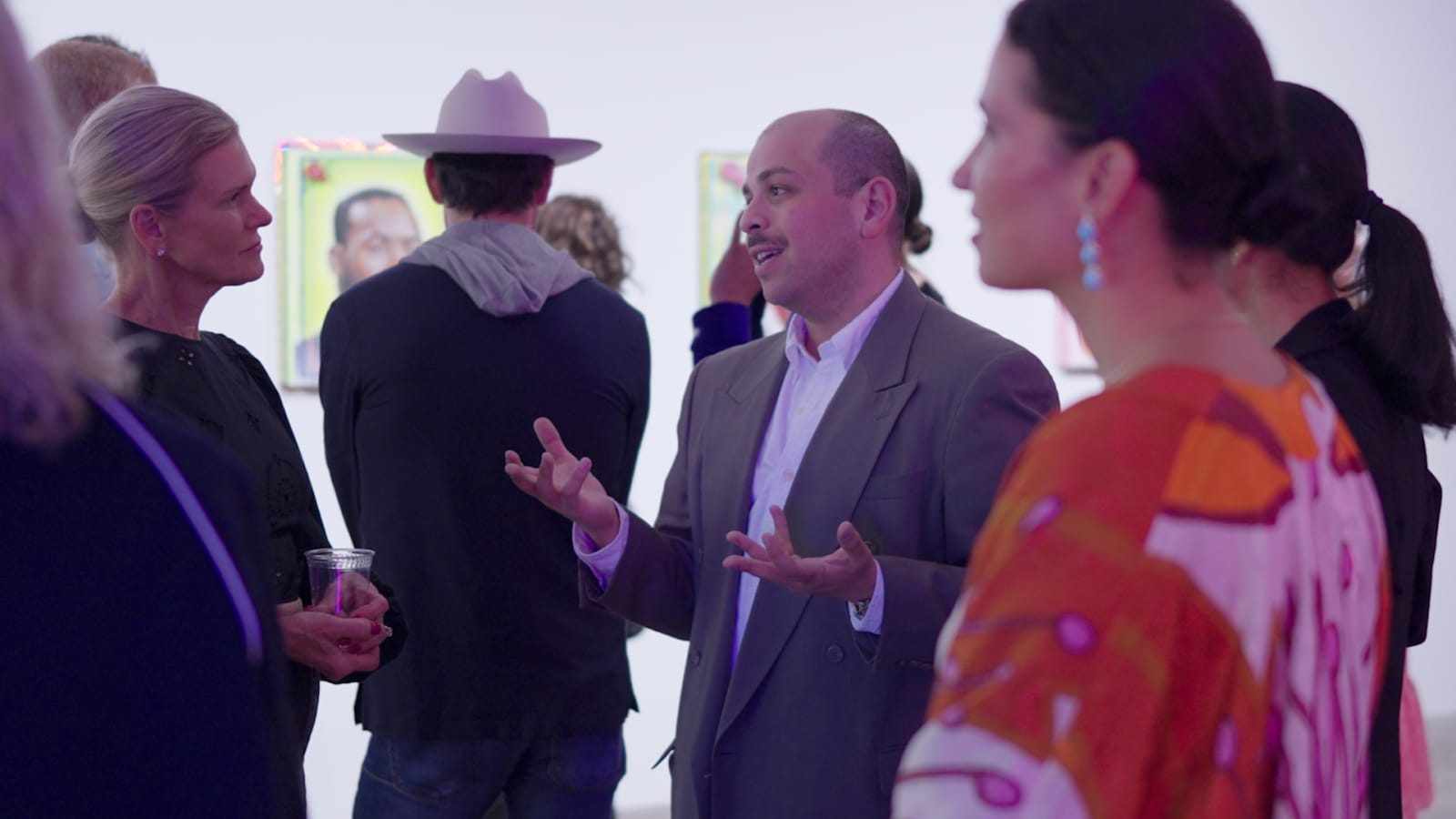 A small group of gallery guests in quiet conversation, framed portrait paintings visible on the wall behind them.