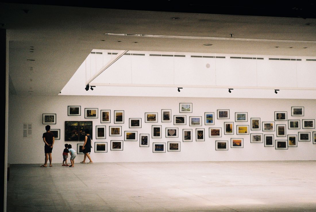 Visitors viewed from a distance walking past a gallery wall densely hung with small framed artworks