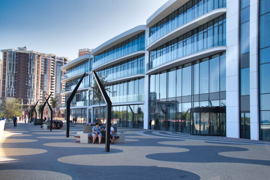 A curved glass office building with a landscaped plaza in front, pedestrians seated and walking among tall abstract black sculptural posts