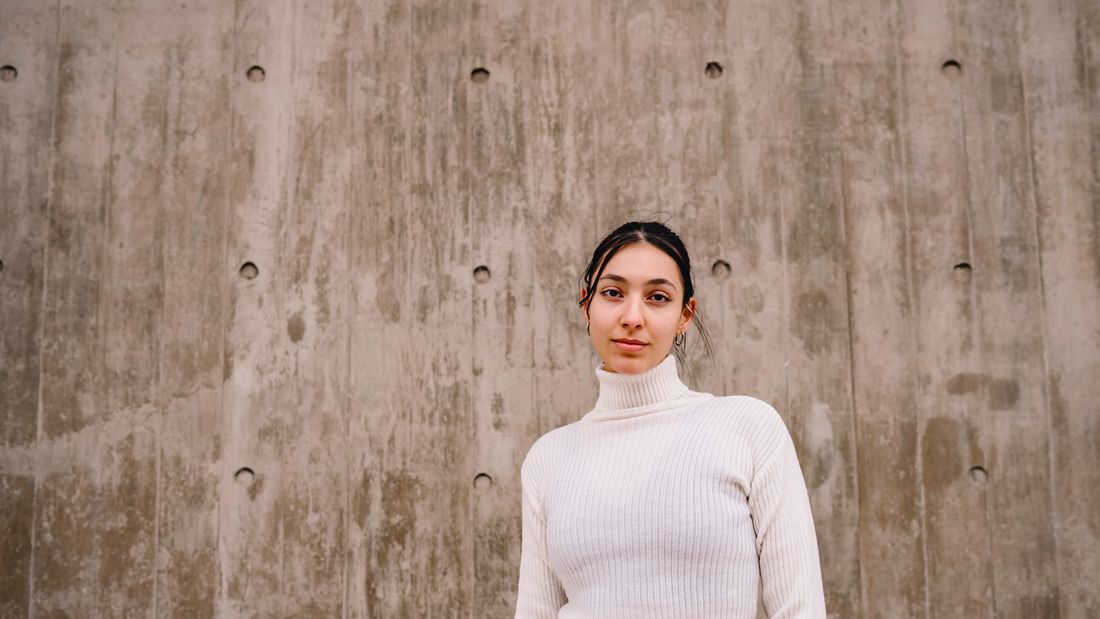 A calm three-quarter portrait of a young woman in a white sweater standing against a plain neutral concrete wall in natural light.