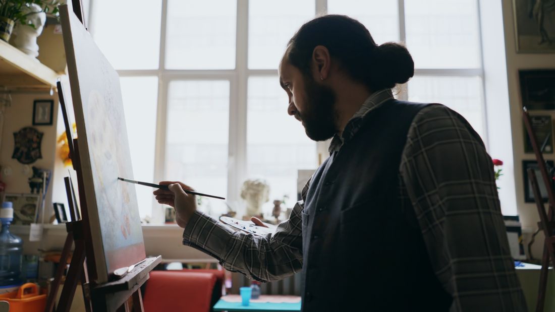 An artist working at an easel in a quiet, sunlit studio, seen in profile