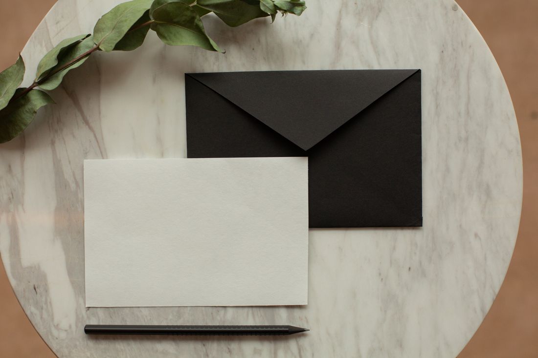 Overhead flat lay of a blank white card, black envelope, and pencil on a marble table with a dried sprig