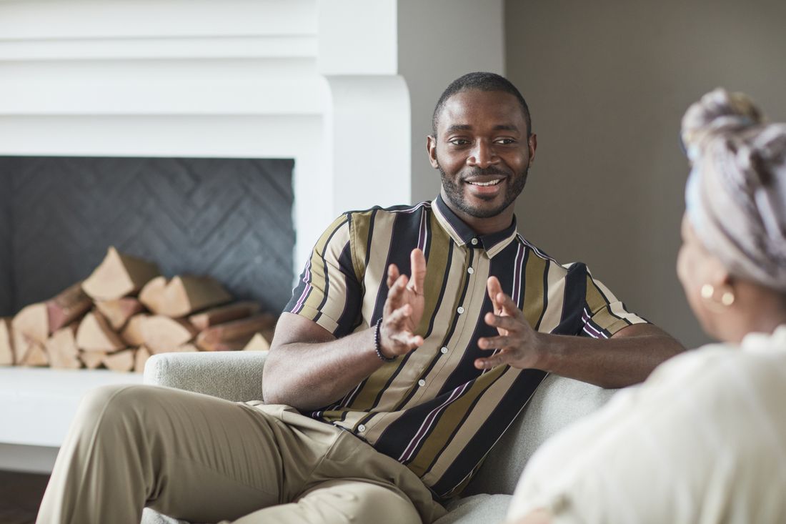 A man seated on a couch, smiling and gesturing mid-sentence while talking to someone off-frame