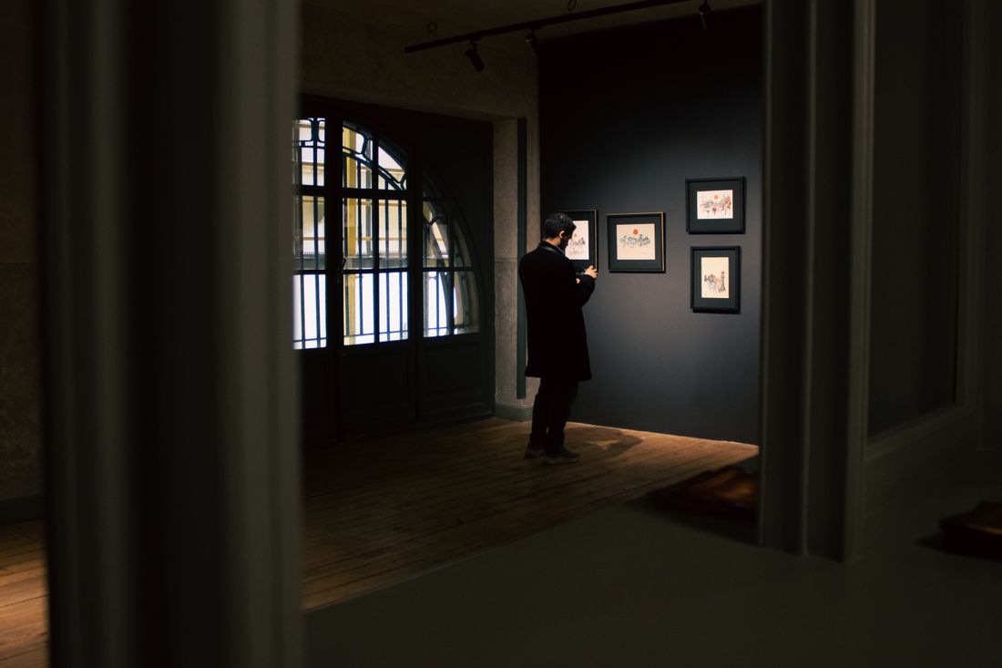 A visitor standing quietly before a dark gallery wall hung with small framed illustrations, seen through an open doorway