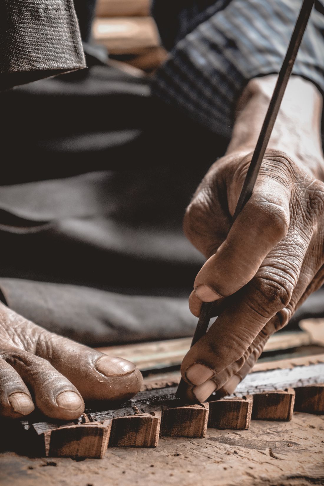Close-up of a craftsperson's weathered hands working a chisel against small wooden pieces on a worn table, no face in frame