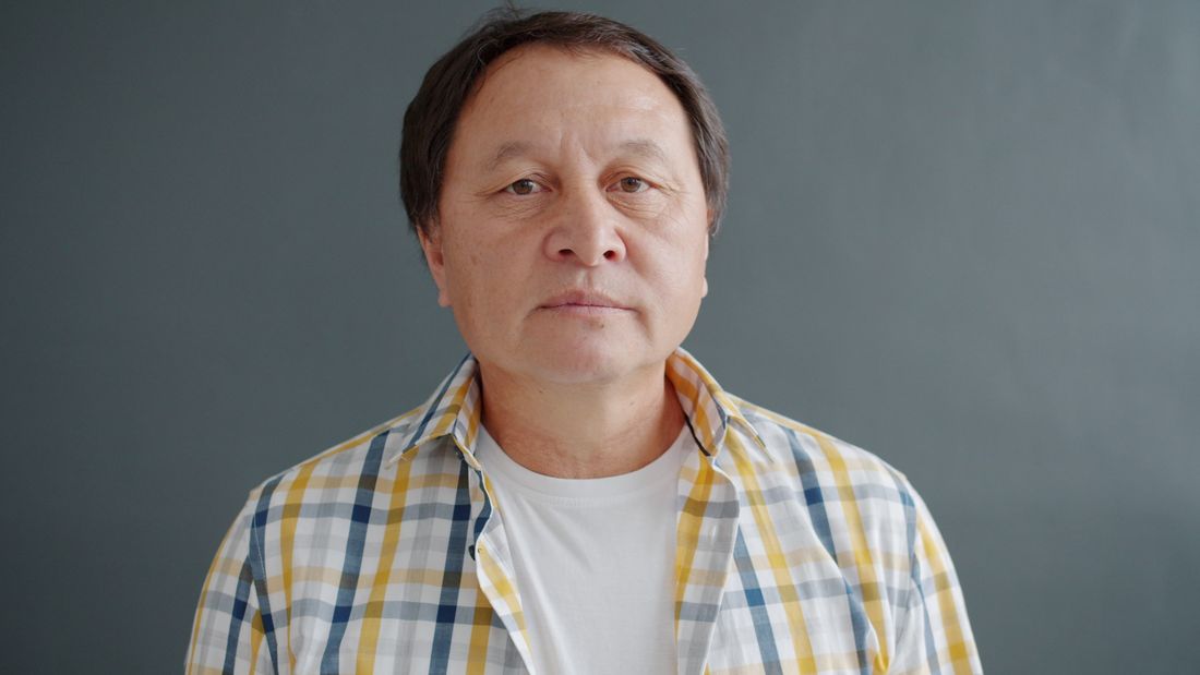 A middle-aged person in a plaid shirt standing calmly against a plain gray wall