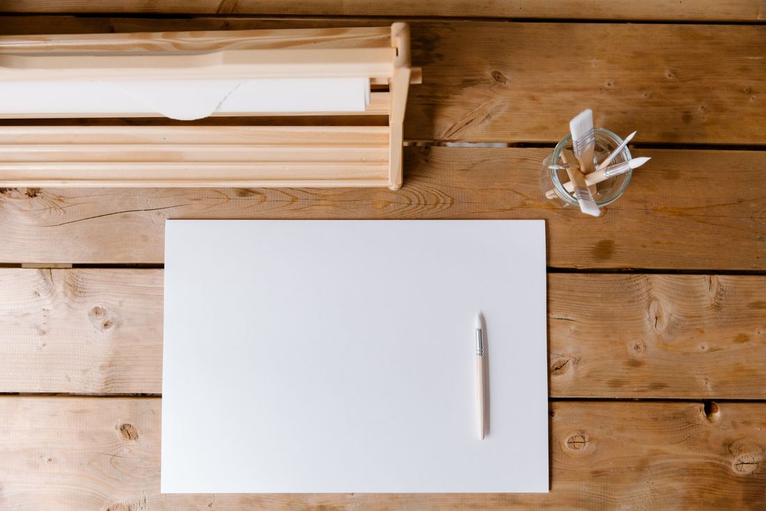 A blank sheet of paper and a plain wooden pen on a bare wooden work table, with brushes in a glass jar nearby