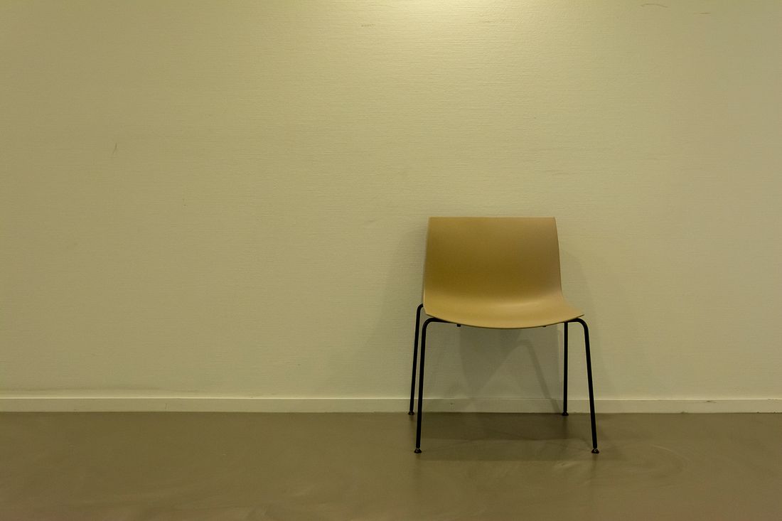 A single simple chair standing against a plain, warmly lit wall in an otherwise empty room.