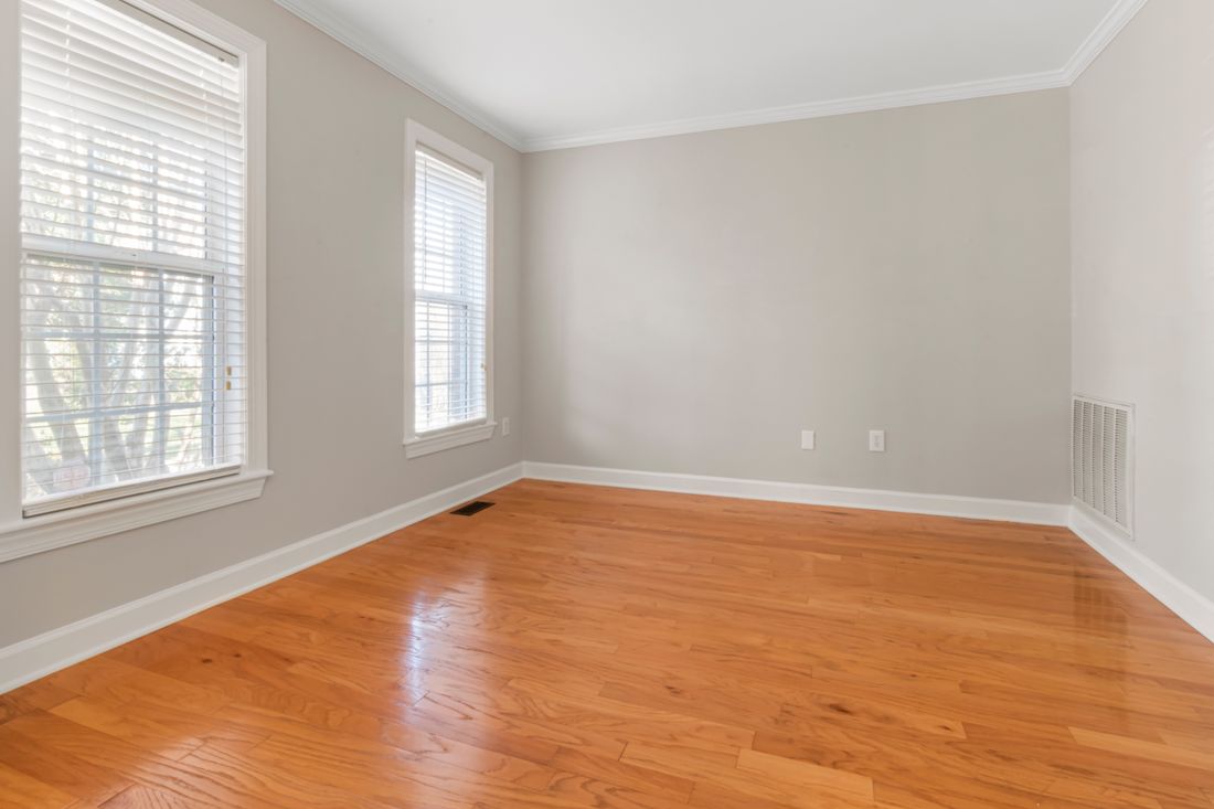 An empty room with pale gray walls, two bright windows, and a polished hardwood floor in natural daylight.
