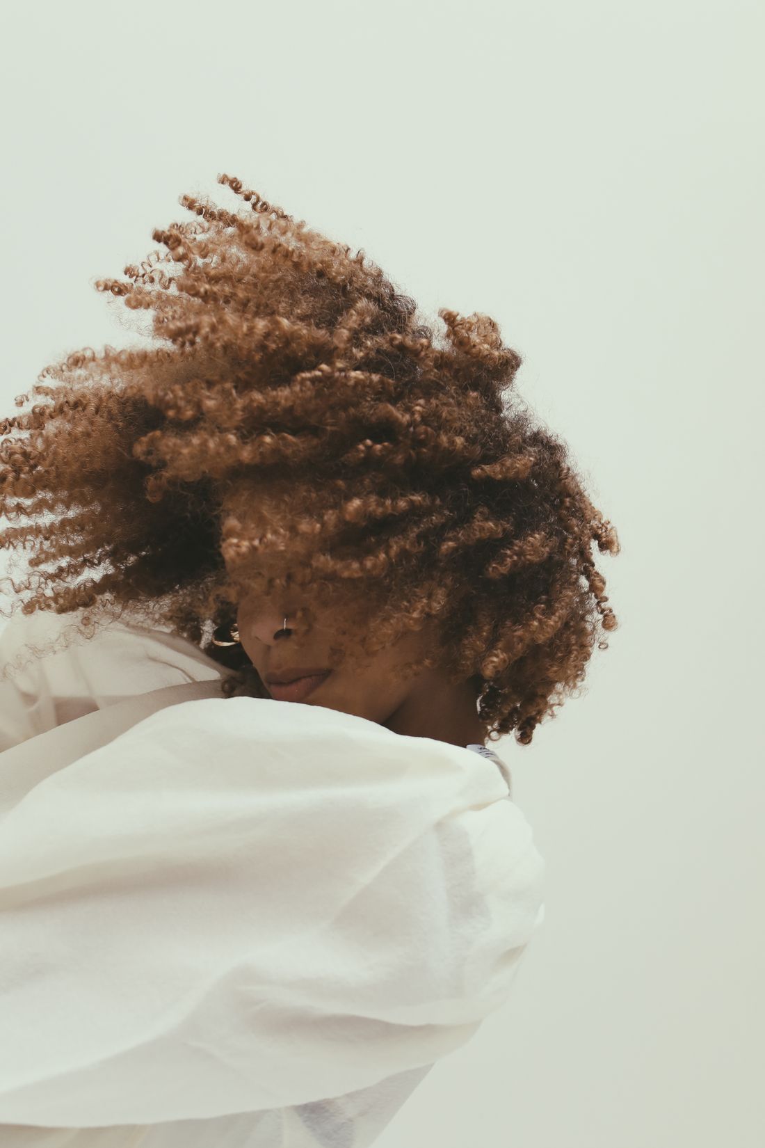 A woman's curly hair caught mid-motion against a plain light background, her face obscured, one arm raised in a white sleeve