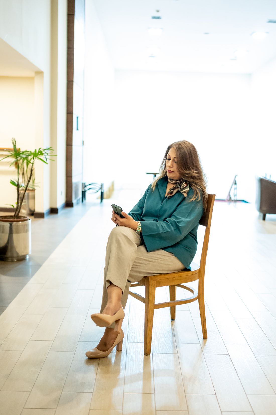 A woman in business attire seated on a chair in a bright office lobby, checking her phone while waiting