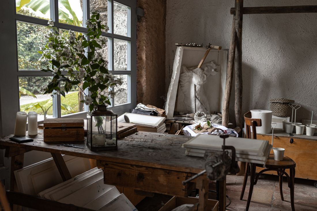 A cluttered artist studio workbench beneath tall windows, with a plaster relief sculpture propped against the wall in natural daylight