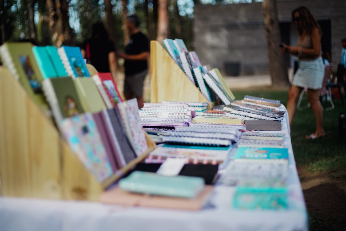 A long table at an outdoor market lined edge to edge with small stacks of colorful notebooks and booklets, shot down the table with people browsing softly out of focus in the background.