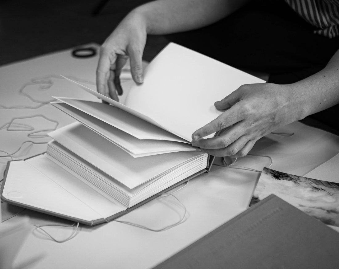 Close black-and-white view of two hands mid-turn on the blank pages of an open handmade booklet resting on a table, with loose paper and string softly out of focus behind it.