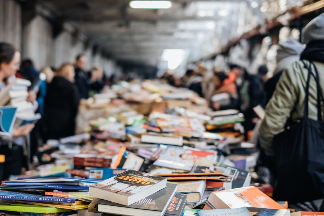 Over-the-shoulder view down a packed indoor book market aisle, back and shoulder of a visitor in the foreground beside a long table piled with stacks of paperback and hardcover books, other browsers packed close together further down the aisle.