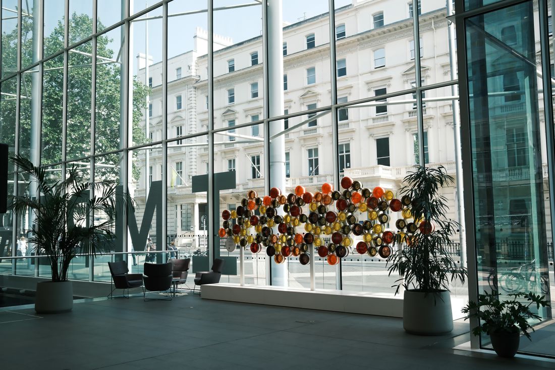 Modern office lobby with a floor-to-ceiling glass curtain wall and a large multicolored sculptural art installation, with seating and potted plants nearby