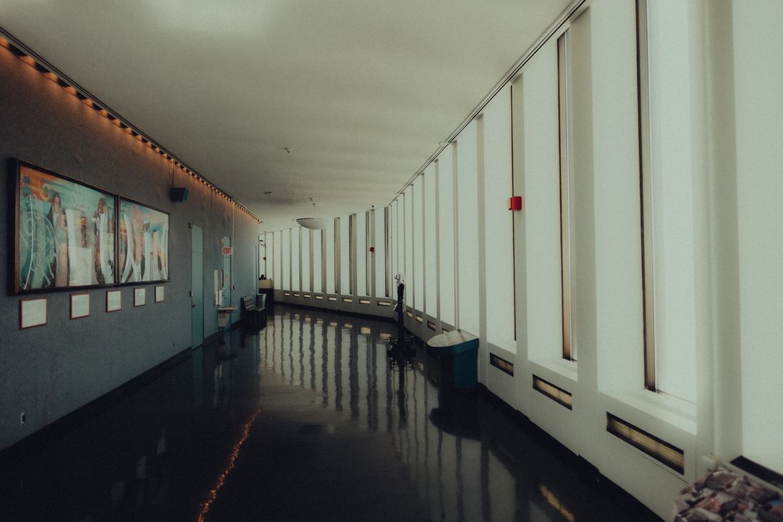 Modern building corridor with framed artwork on a side wall and a glossy polished floor reflecting the ceiling and windows