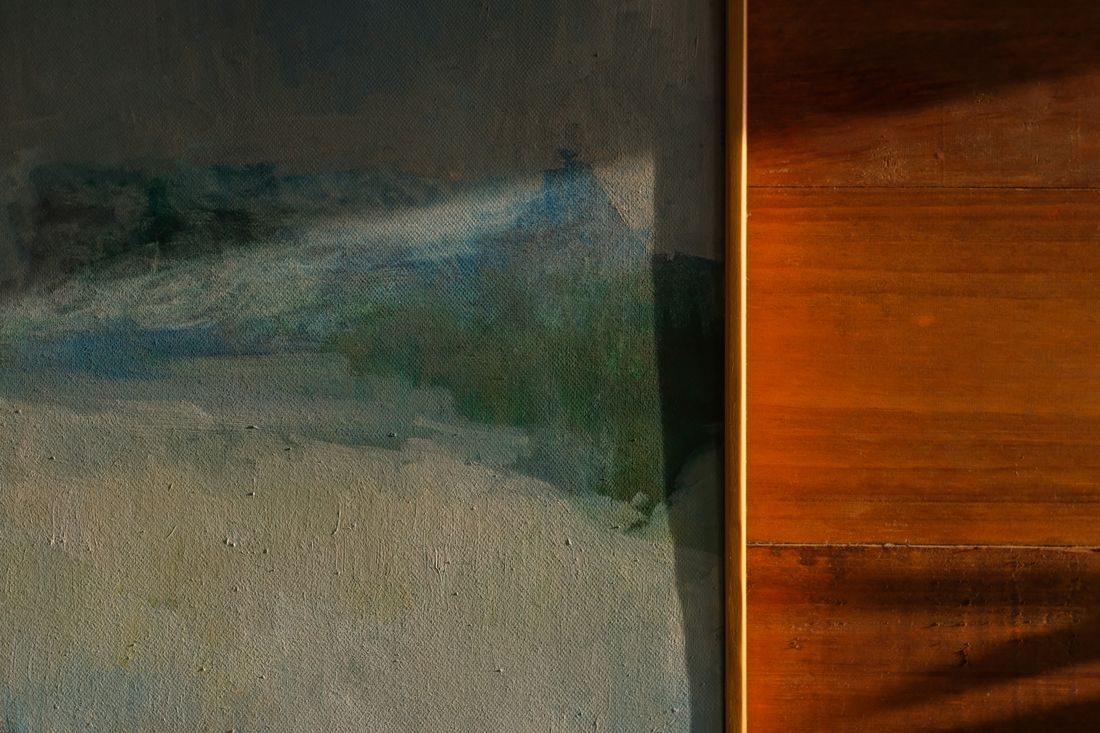 A close, warmly lit view of a finished painting's edge beside a wood-toned wall surface, showing texture and directional light.