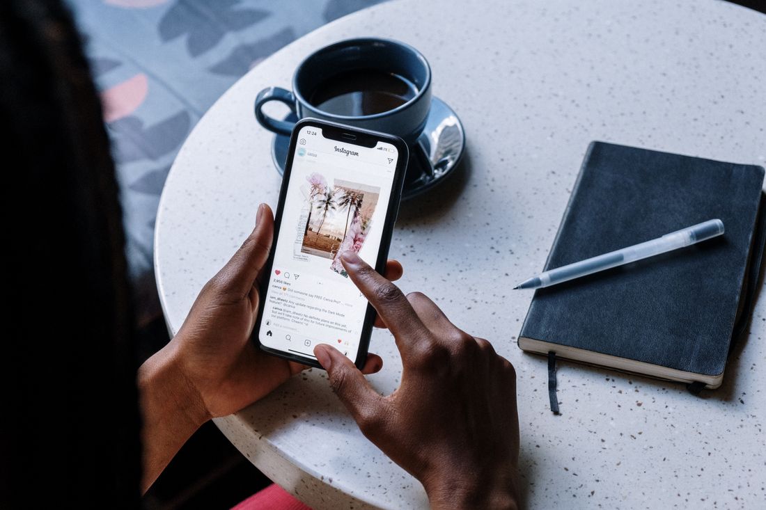 Hands holding a phone showing an image feed on a small table beside a coffee cup and notebook