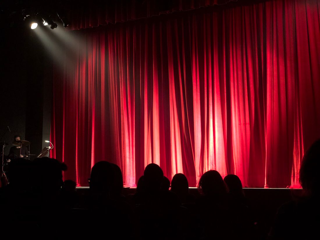 Audience silhouettes facing a lit stage with red curtains, waiting for a program to begin