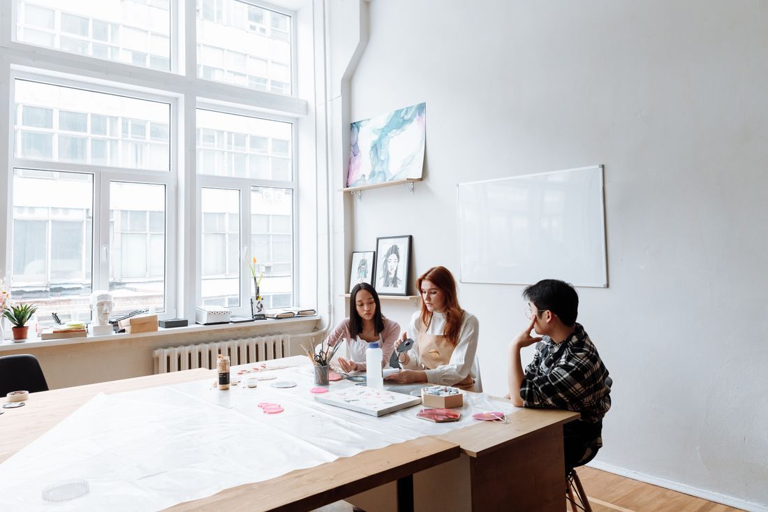 Three adults gathered around a table in a bright studio with a large window, working with craft materials, hands and shoulders visible, a blank whiteboard on the wall behind them.