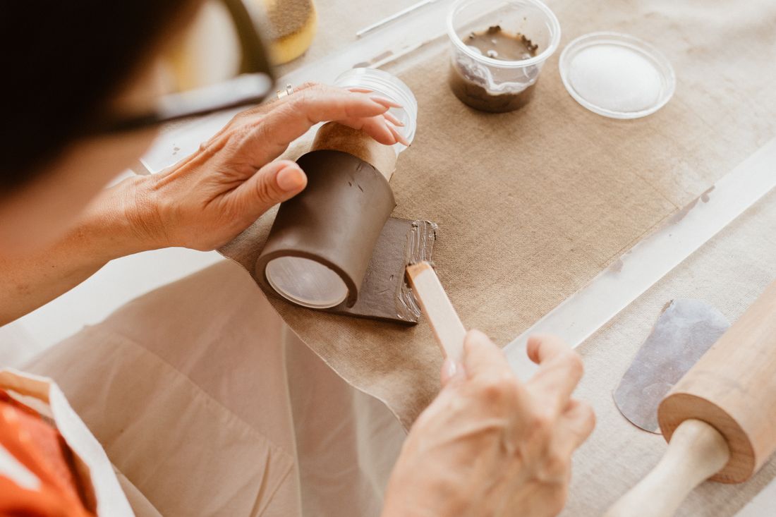 Close overhead view of an adult's hands shaping a piece of clay with a wooden tool at a work table, materials scattered nearby, no face visible.