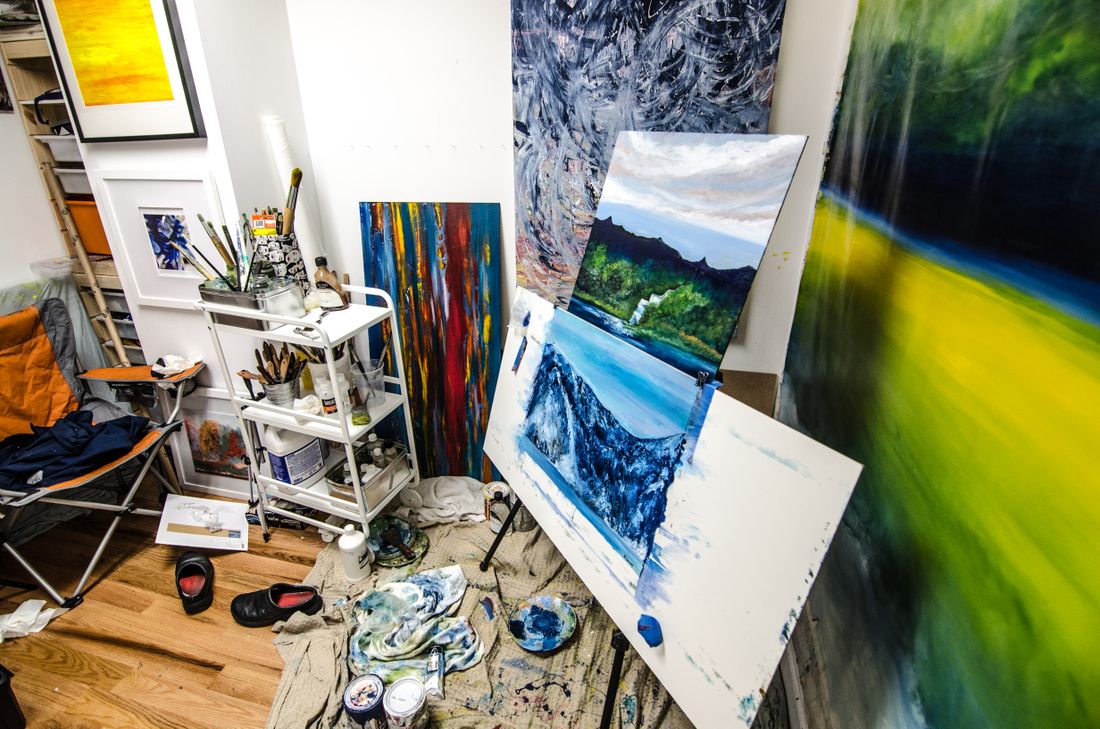 A working artist studio corner with finished paintings leaning against the wall and on easels, a supply cart beside them