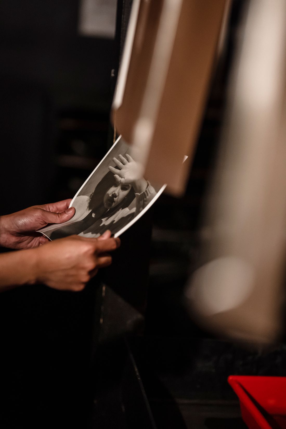Hands holding a freshly printed black-and-white photograph in a dim darkroom, inspecting it against the light