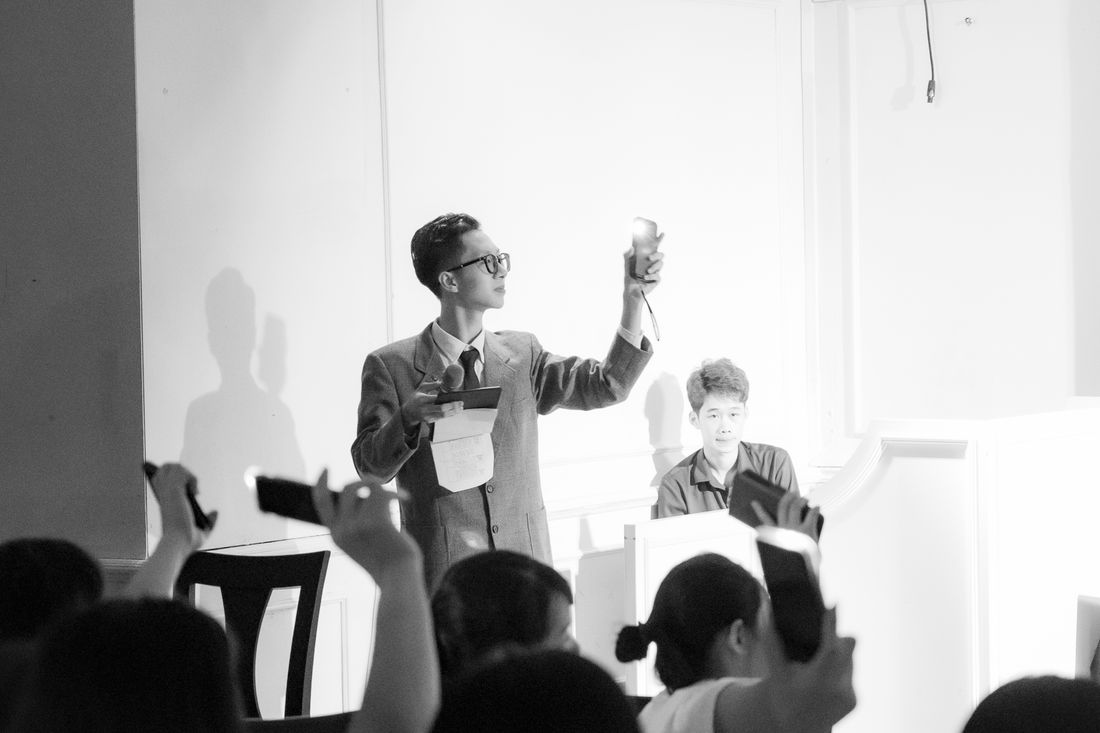 A black-and-white auction scene with a presenter at a podium and bidders raising paddles and phones in the crowd