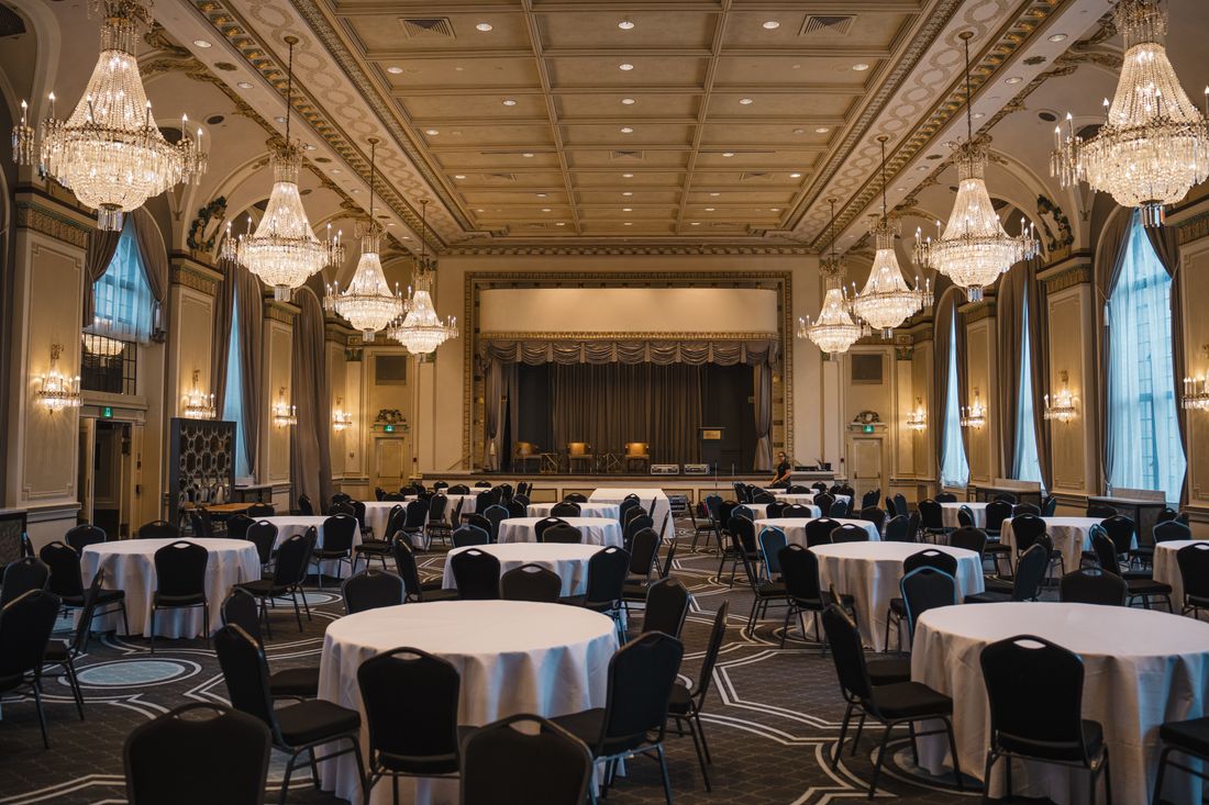 An elegant empty ballroom with round tables, white linens, and crystal chandeliers set for an evening event