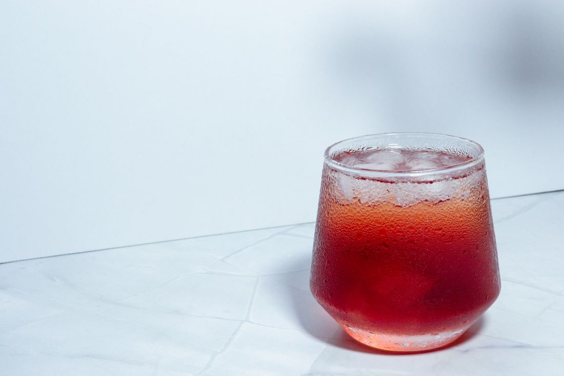 A short glass of a red beverage over ice, condensation beading on the glass, set on a marble surface against a plain light backdrop.