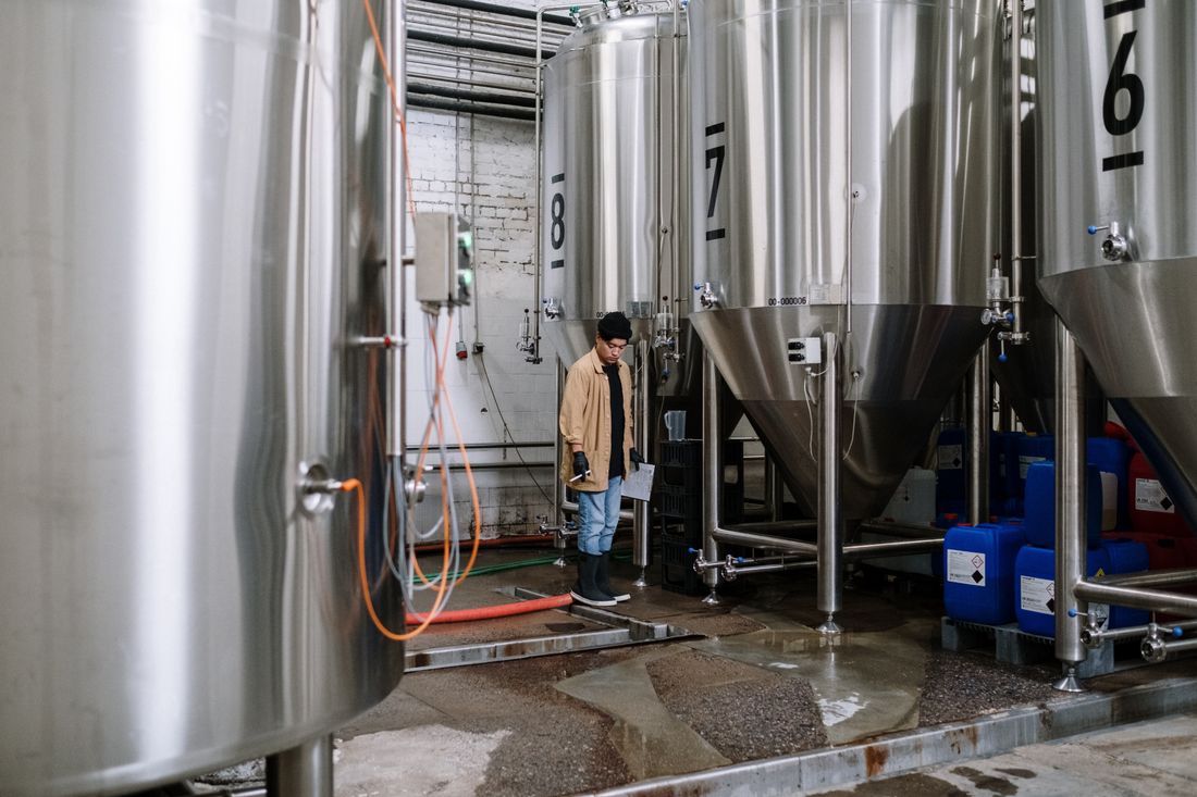A worker in a canvas jacket checking a clipboard while inspecting a row of large stainless steel fermentation tanks in a production facility.
