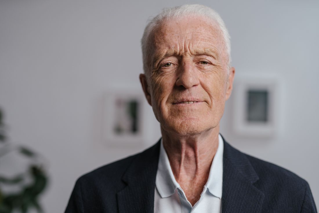An older man in a dark suit jacket stands calmly against a plain wall, with softly blurred framed pictures in the background