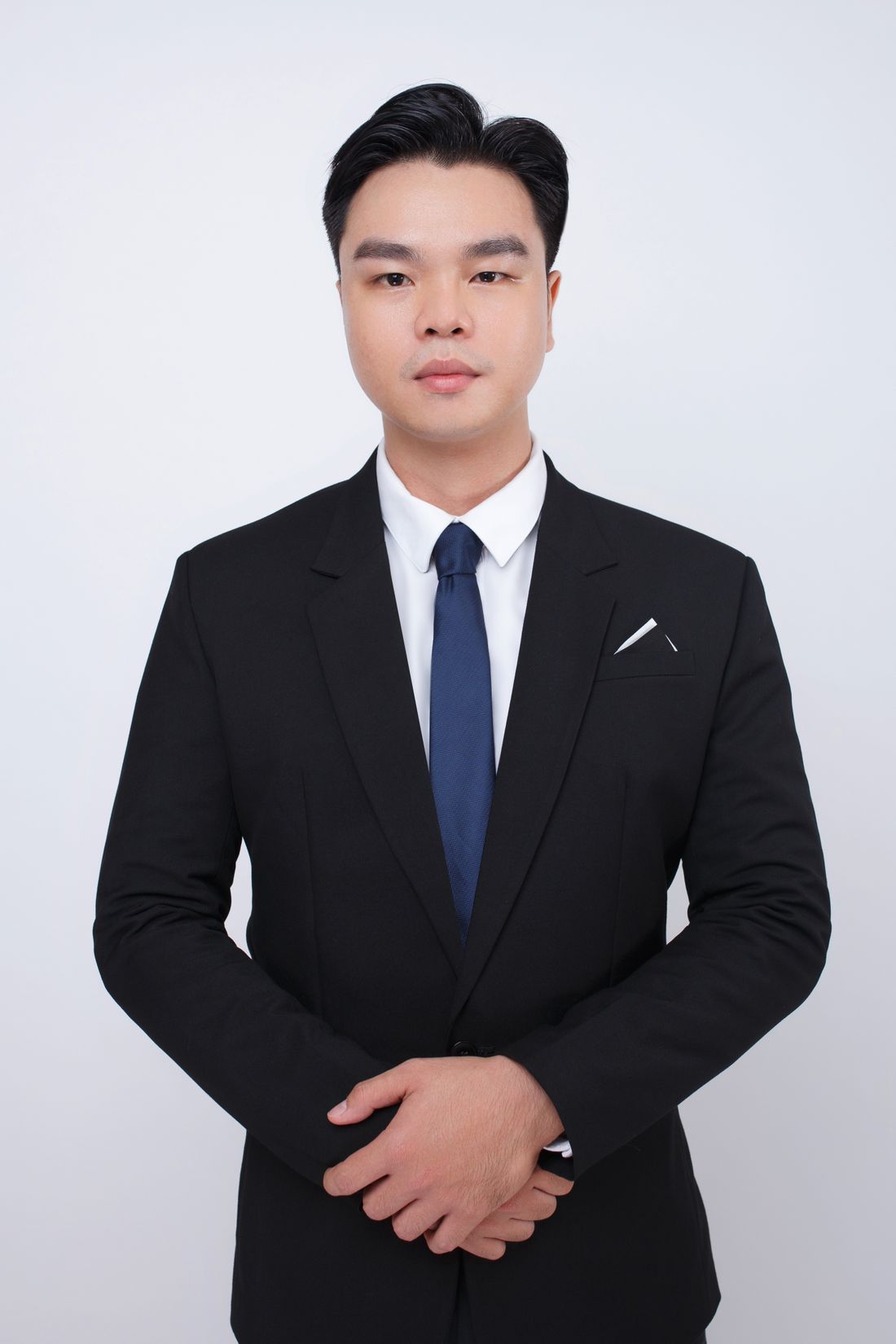 A vertical portrait of a man in a black suit and tie with hands clasped, photographed against a plain white background
