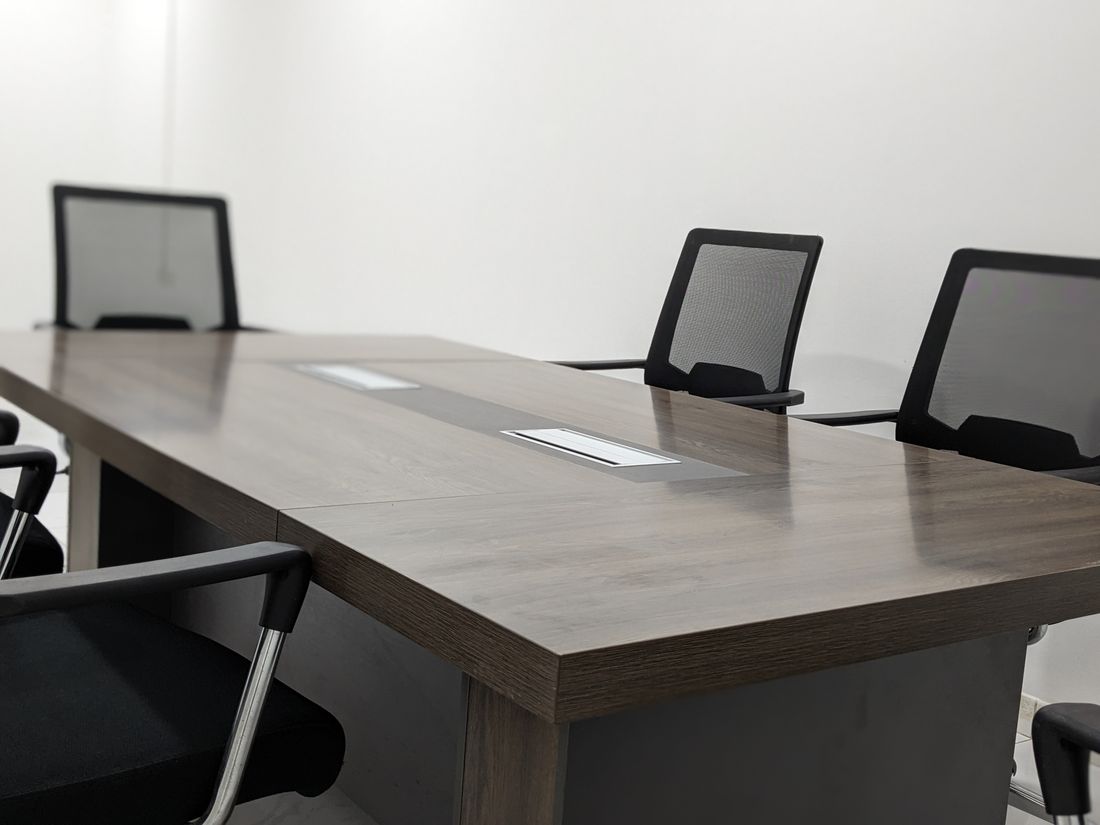 An empty conference room with a long wooden table and dark mesh office chairs against a plain white wall