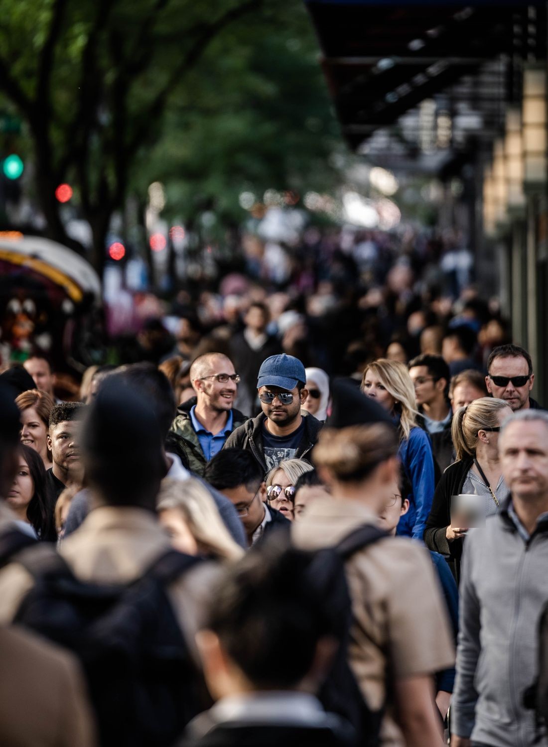 A dense, anonymous crowd of pedestrians filling a city sidewalk beneath trees and storefront awnings