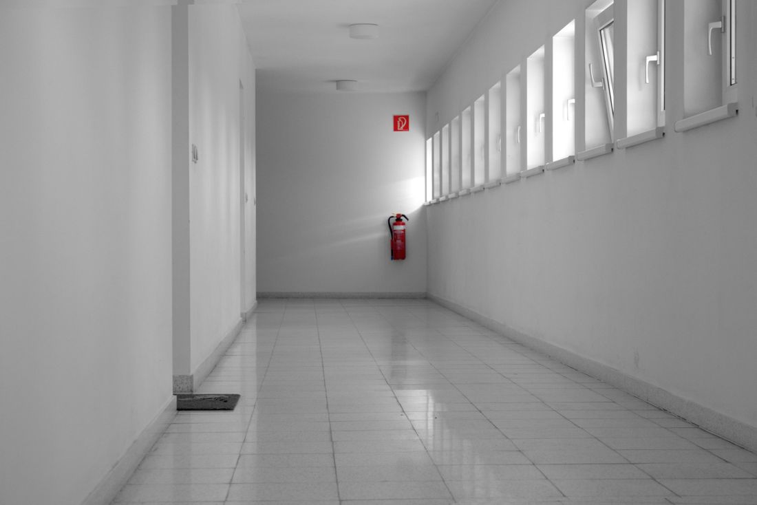 A plain white institutional corridor with a door on one side, a row of small windows on the other, and a red fire extinguisher on the wall