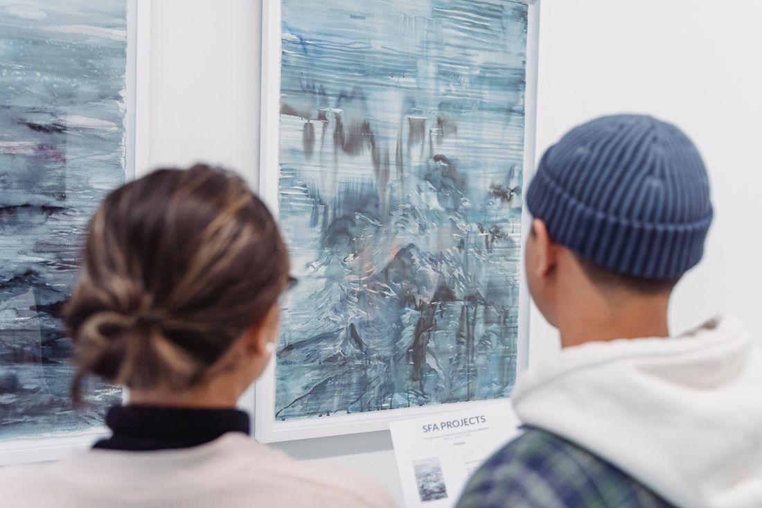 Two people seen from behind standing back and considering large framed paintings on a quiet gallery wall