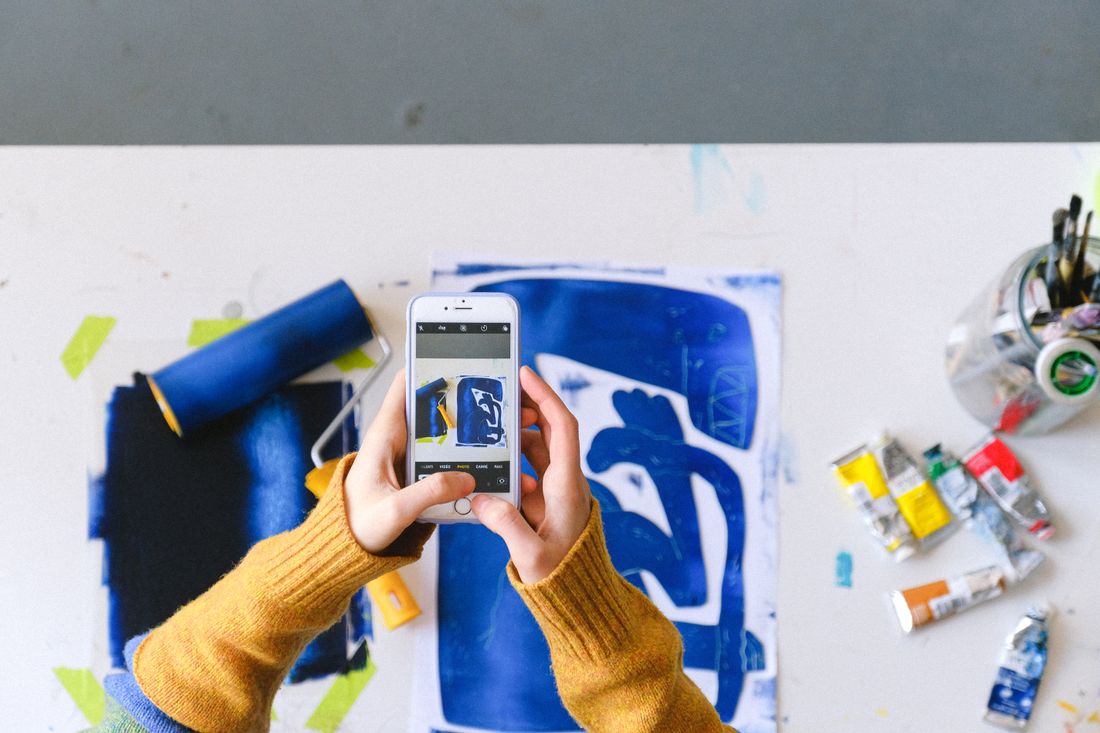 Overhead view of hands holding a phone, photographing a blue and white abstract painting on a table
