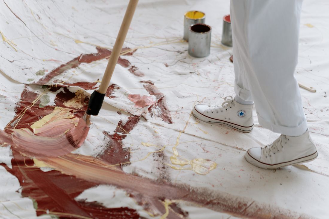 Cropped view of an artist's legs and sneakers mid-brushstroke on a large canvas laid on the studio floor, paint cans nearby