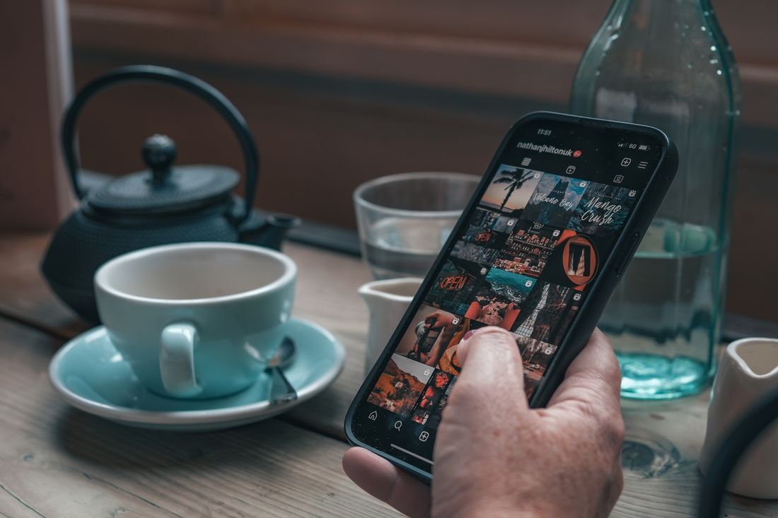 A hand holding a smartphone open to an Instagram photo grid at a table with tea