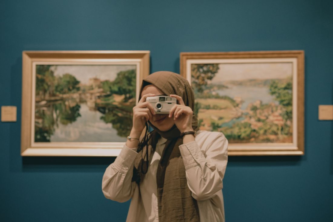 A woman photographing framed landscape paintings on a gallery wall with a camera