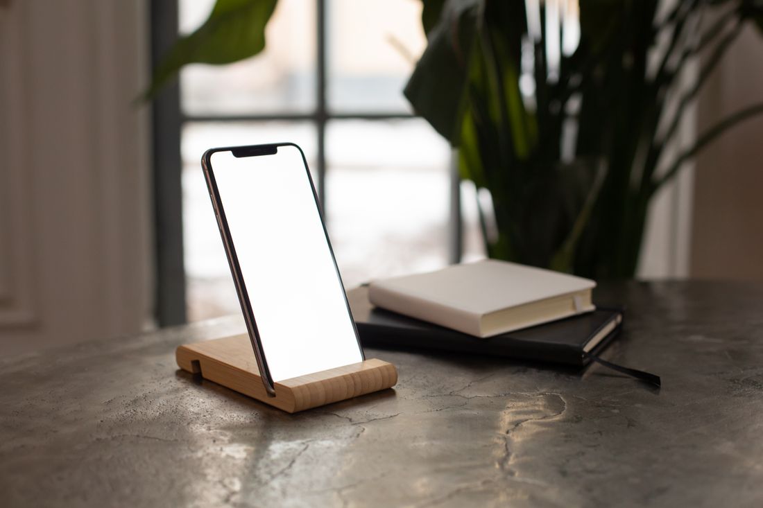 A smartphone propped upright on a wooden stand with a blank screen, beside two closed notebooks on a desk in natural window light.