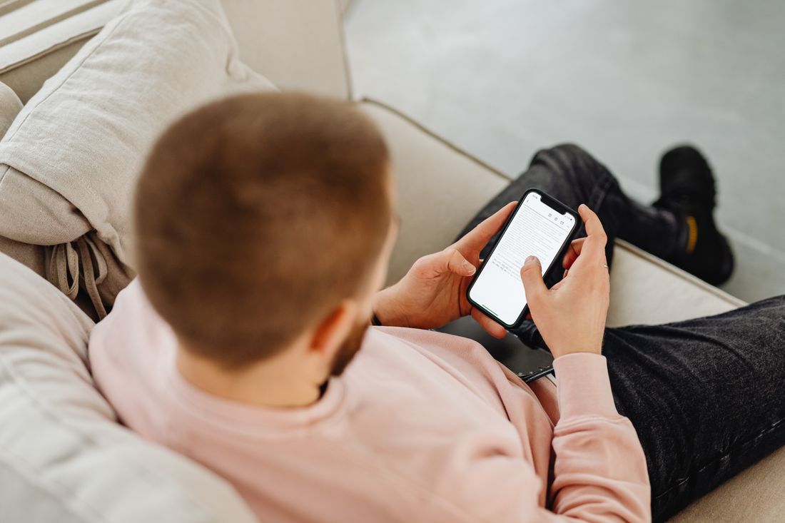 A person viewed over the shoulder, seated on a couch, holding a smartphone with a blank white screen at a slight angle.