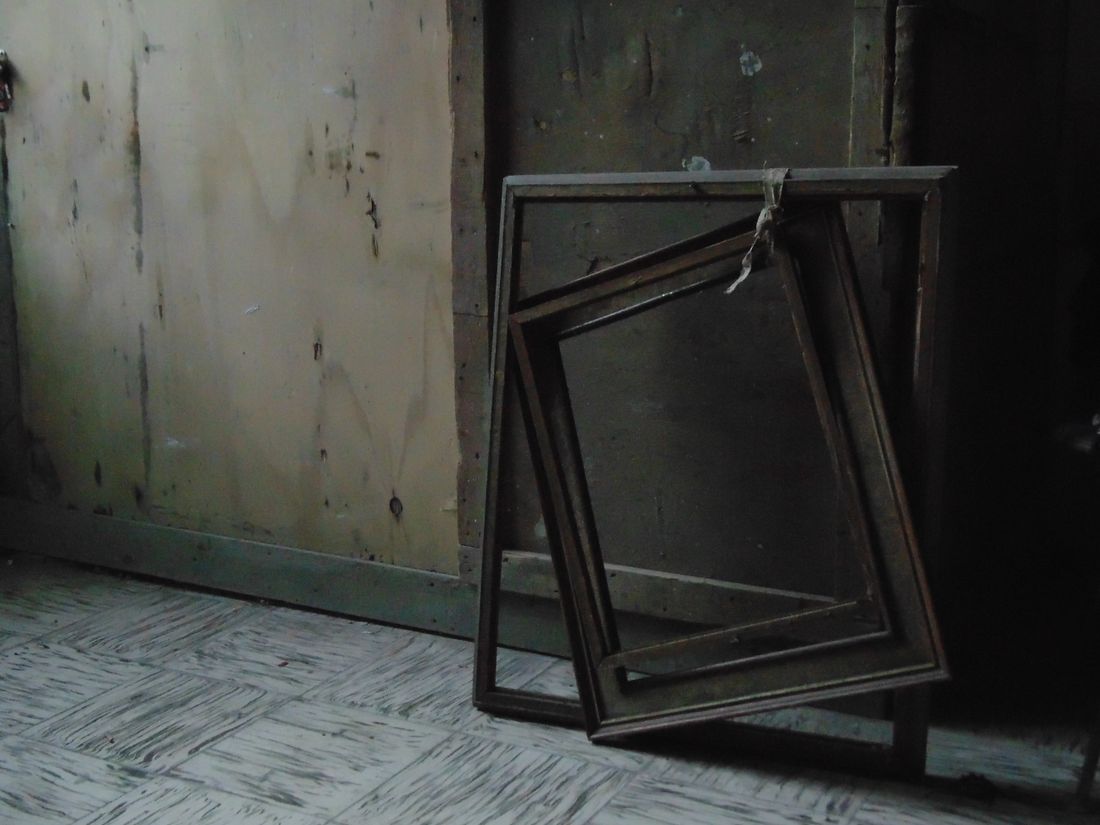 Old wooden picture frames of different sizes leaning together against a faded wall in a dim, unused room